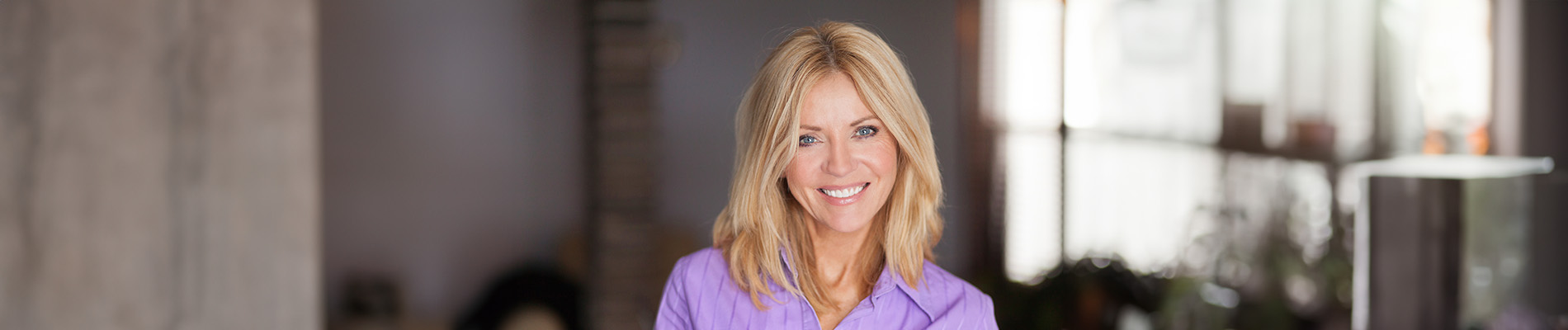 The image is a photograph of a woman with light skin, smiling at the camera. She appears to be in her late twenties or early thirties and has long hair. Her eyes are looking directly at the camera, and she is holding up her index finger near her mouth as if she s making a point or emphasizing something. The background is plain and light-colored, which suggests that this could be a stock photo used for various purposes such as advertising, personal branding, or lifestyle content.